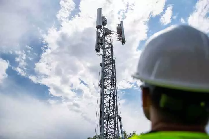 Worker looking up at 5G tower against a cloudy sky.